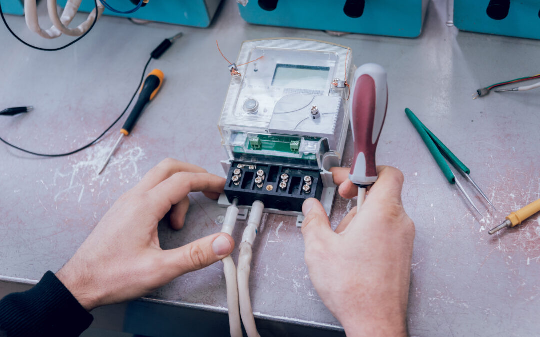 Hands repairing an electrical device on a workbench. The person is working on a digital meter, using a screwdriver while holding wires connected to the terminal. Various tools, including pliers and screwdrivers, are scattered on the surface nearby, indicating an electrical repair process.