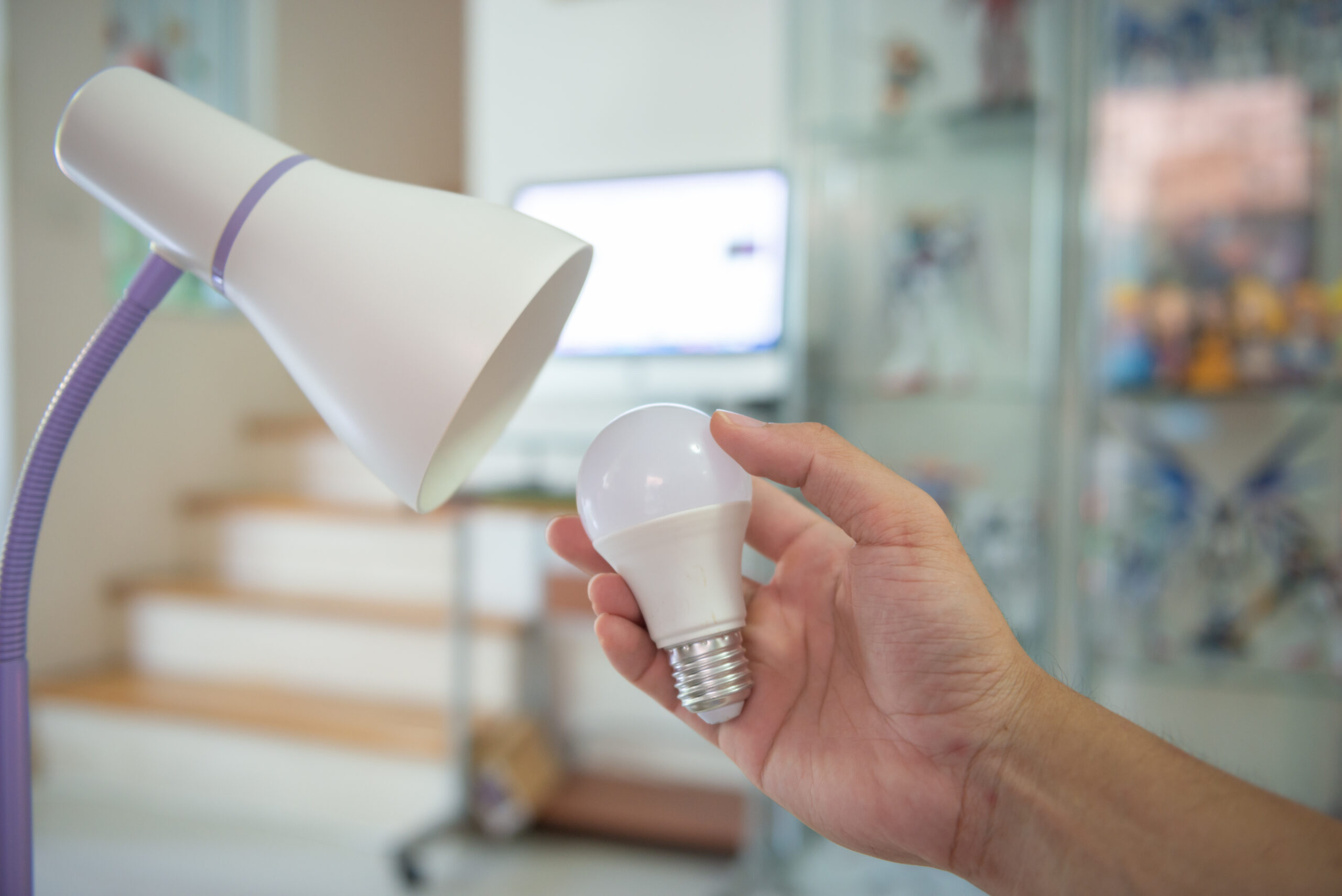 A hand holding an LED light bulb next to a desk lamp, preparing to install or replace the bulb. The background features a blurred interior with shelves and a computer, indicating a home or office setting. The lamp is white with a purple base, adding a touch of modern design to the scene.energy efficient LED lighting