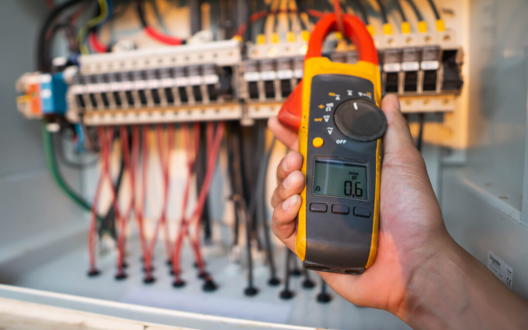 A hand holding a digital clamp meter measuring electrical current in a circuit breaker panel with multiple red and black wires. The device displays "0.6 Amps DC" on its screen. The background showcases the detailed wiring setup of the panel, emphasizing precision and safety in electrical diagnostics and maintenance.